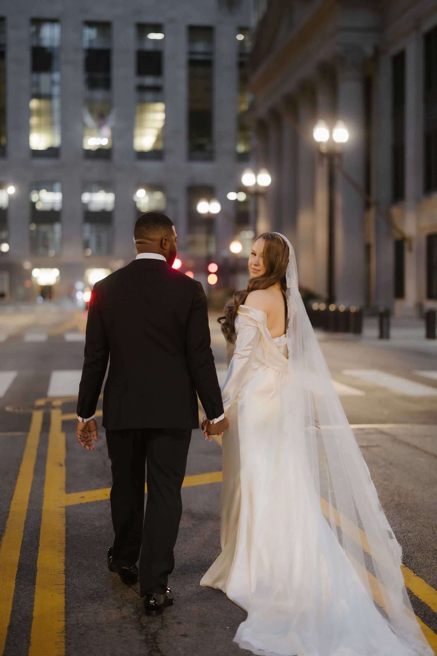 Bride and groom holding hands walking on a city street at night, bride wearing a white gown with veil.