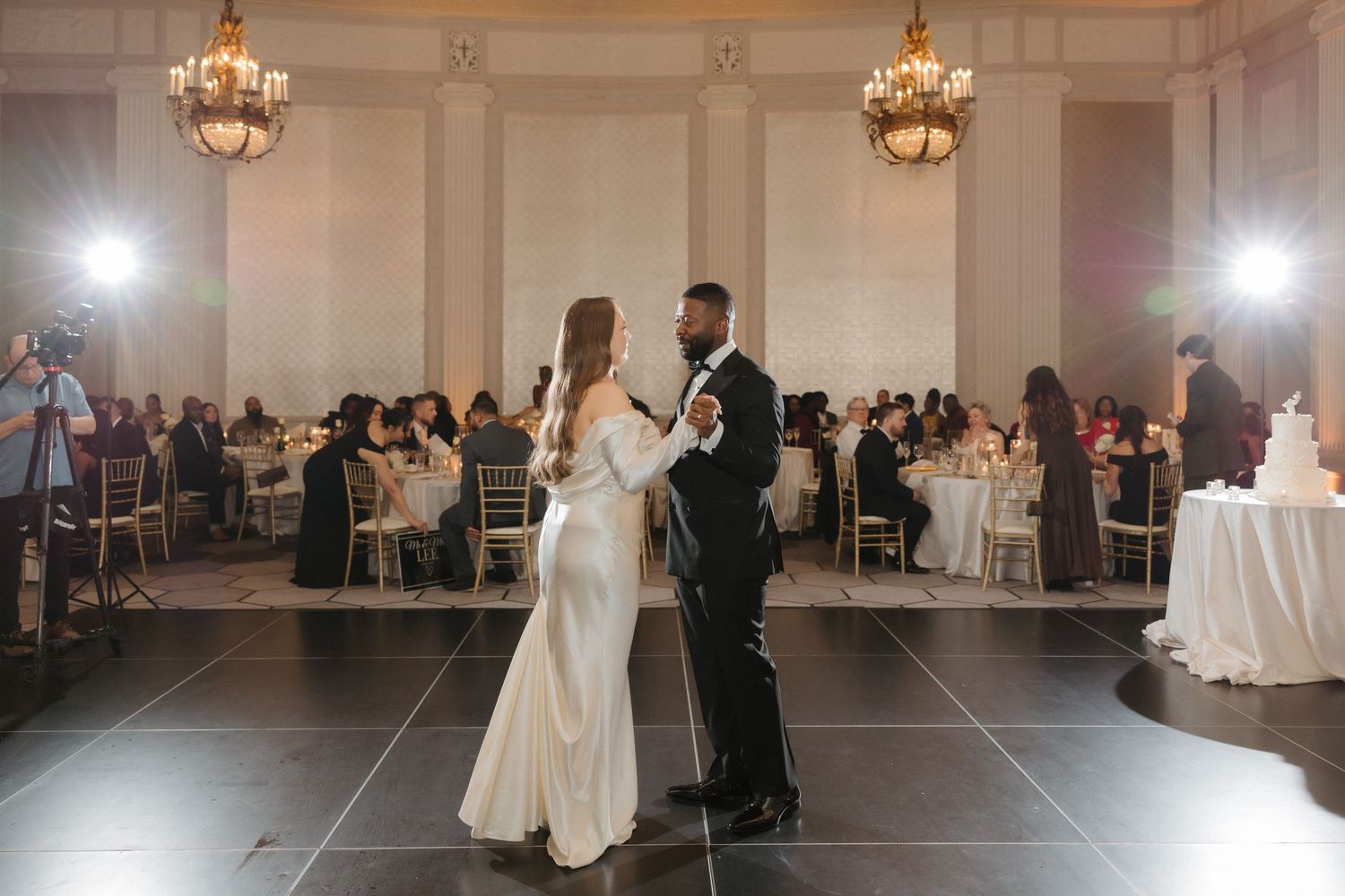 Bride and groom share first dance in elegant ballroom with chandeliers and seated wedding guests in background.