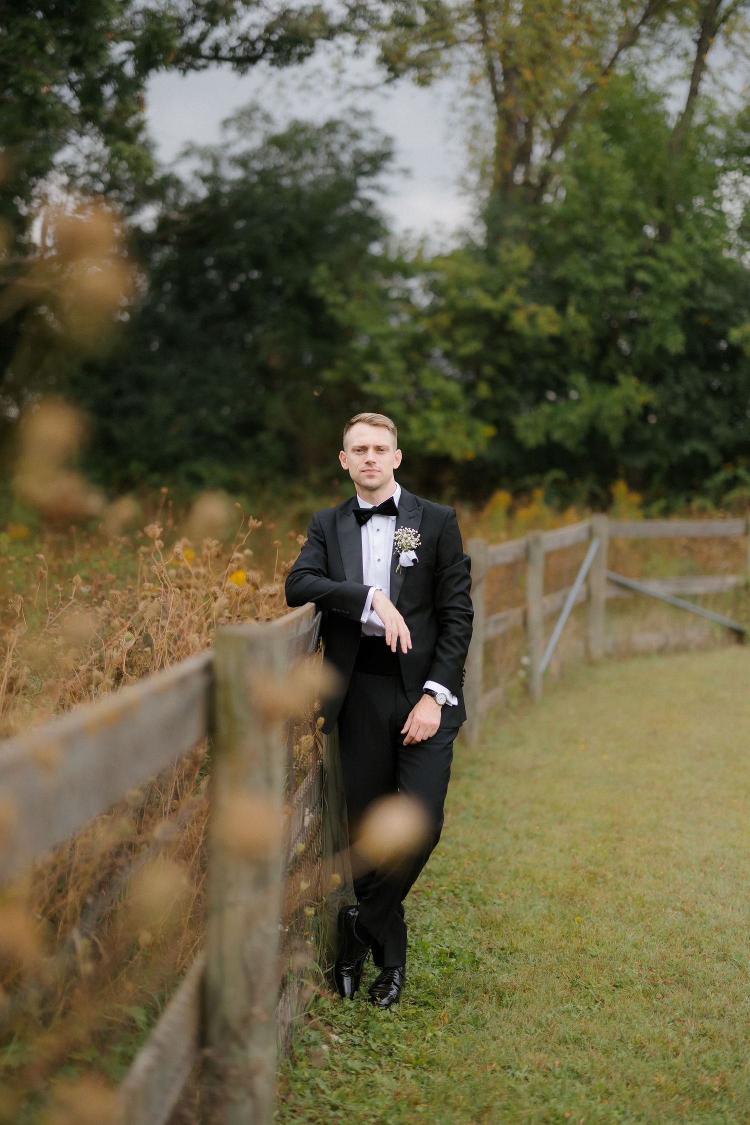 Groom in black tuxedo leaning against wooden fence in lush green outdoor wedding setting.