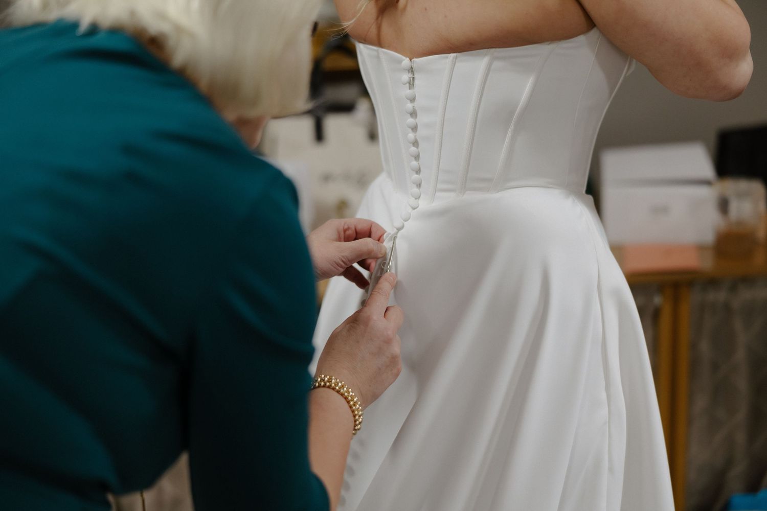 A seamstress fastening buttons on the back of a white bridal gown during a wedding dress fitting.