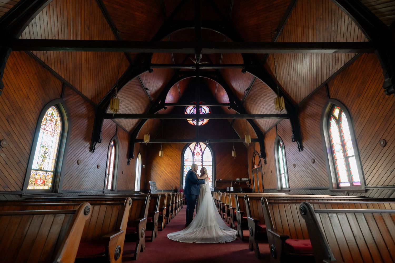 Bride and groom stand at the altar of a stunning wooden church with stained glass windows during their wedding ceremony.