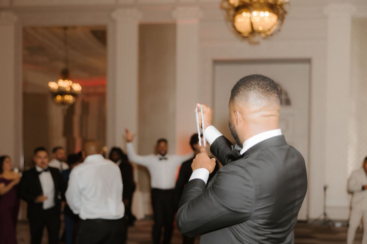 Groom in black tuxedo photographing wedding reception guests celebrating on dance floor in elegant ballroom.