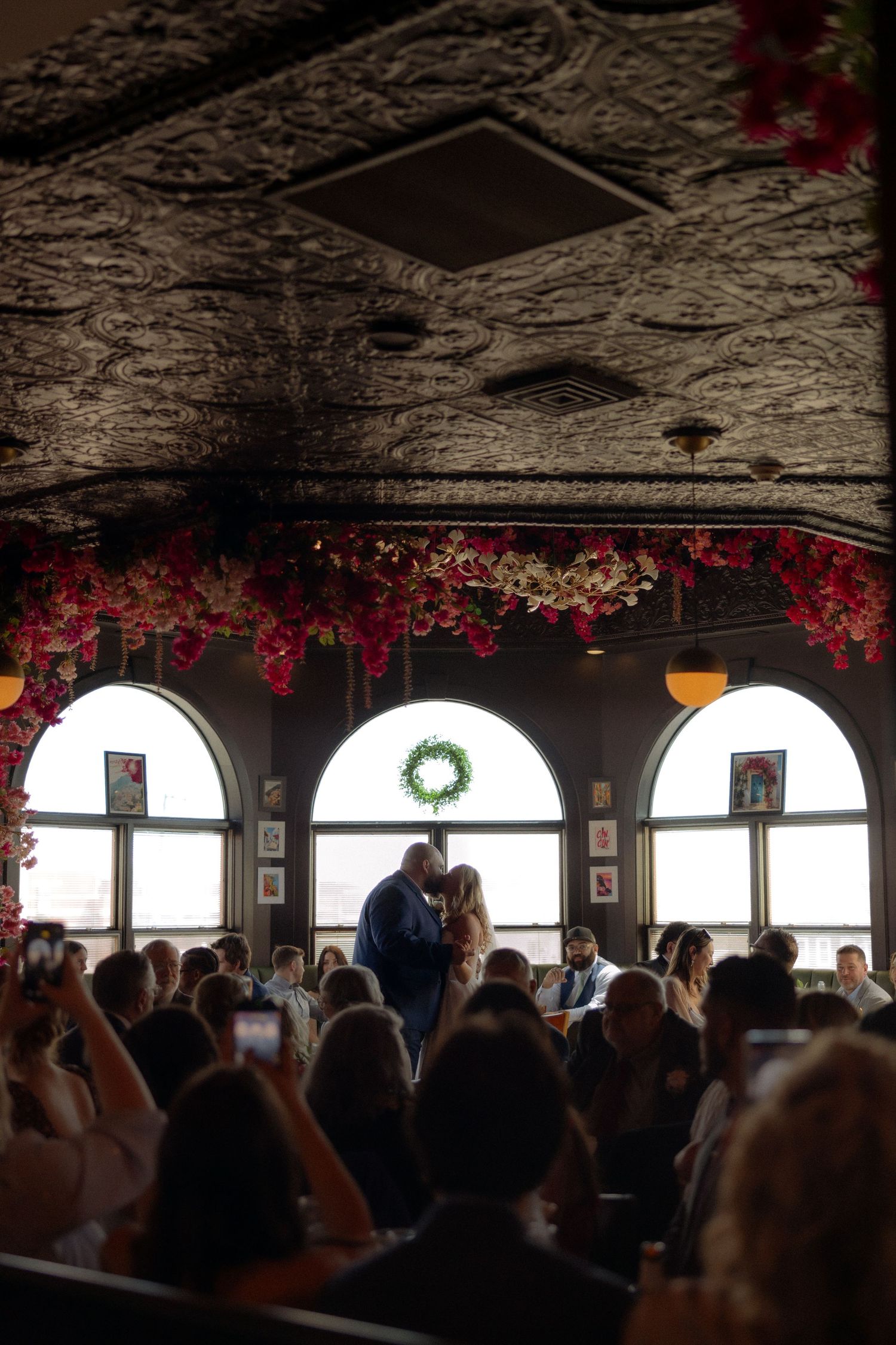 Couple shares a kiss at wedding ceremony in elegant venue with floral arch and arched windows.