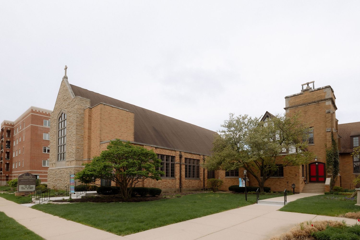 Historic brick church with Gothic architecture, green lawn, and tower under overcast skies in a suburban setting.