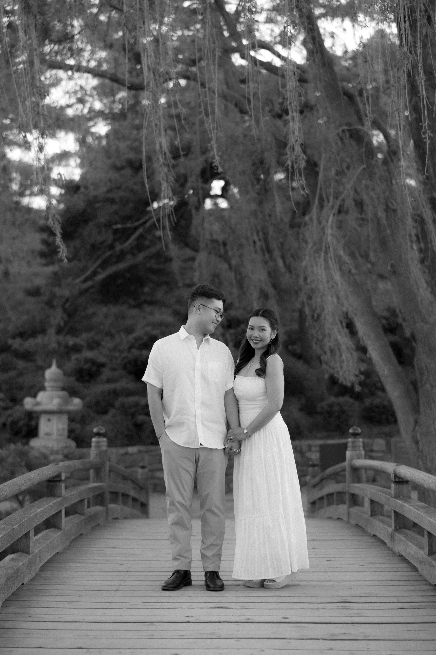 Black and white photo of a couple holding hands on a wooden bridge surrounded by lush trees.