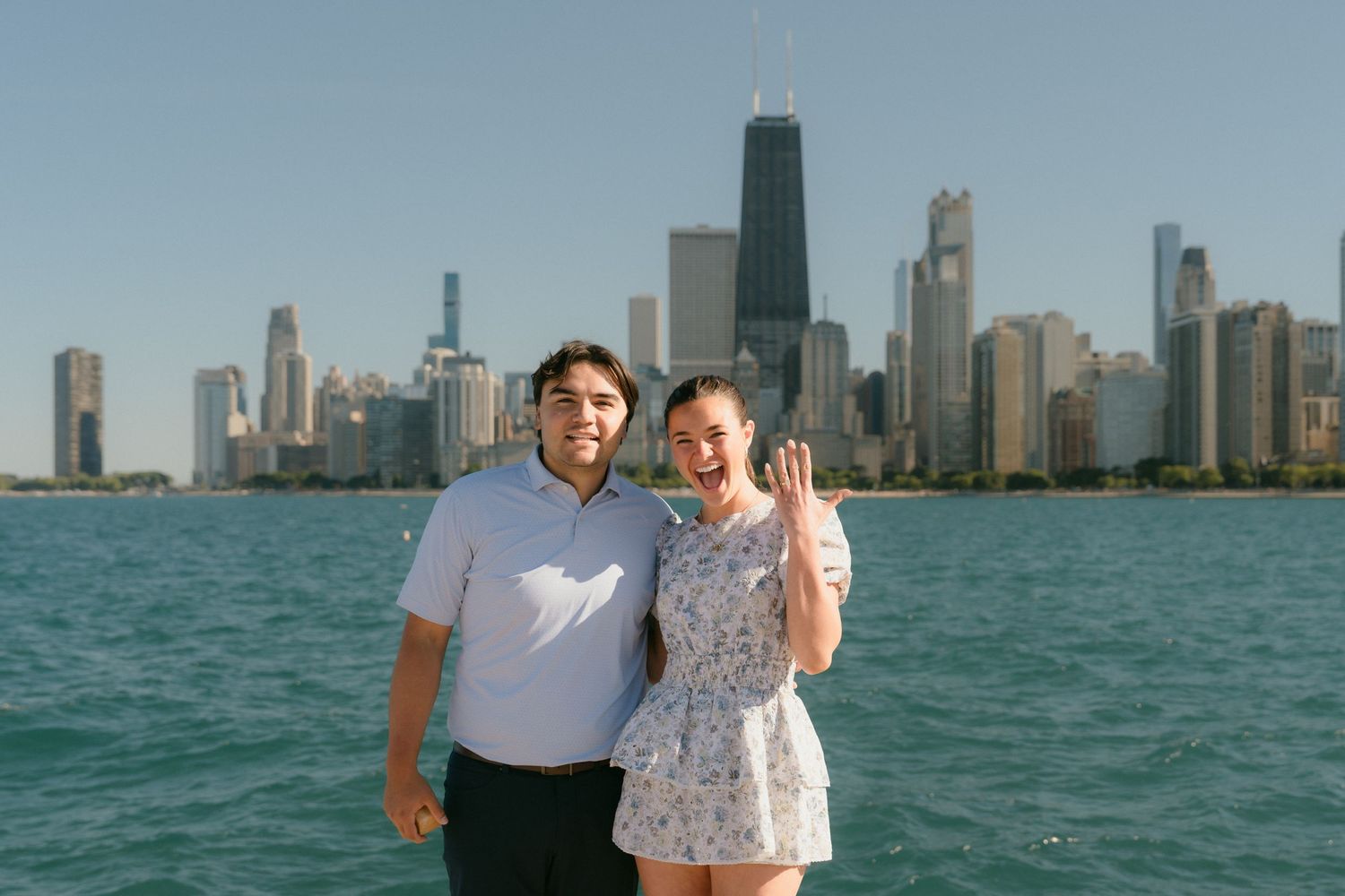 Newly engaged couple smiling by Lake Michigan with Chicago skyline, woman showing off engagement ring.