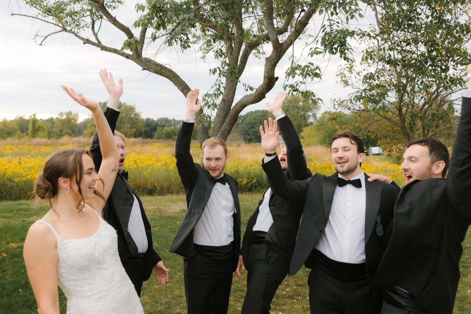 Bride and groomsmen in tuxedos celebrating outdoors with raised hands near a wildflower field.