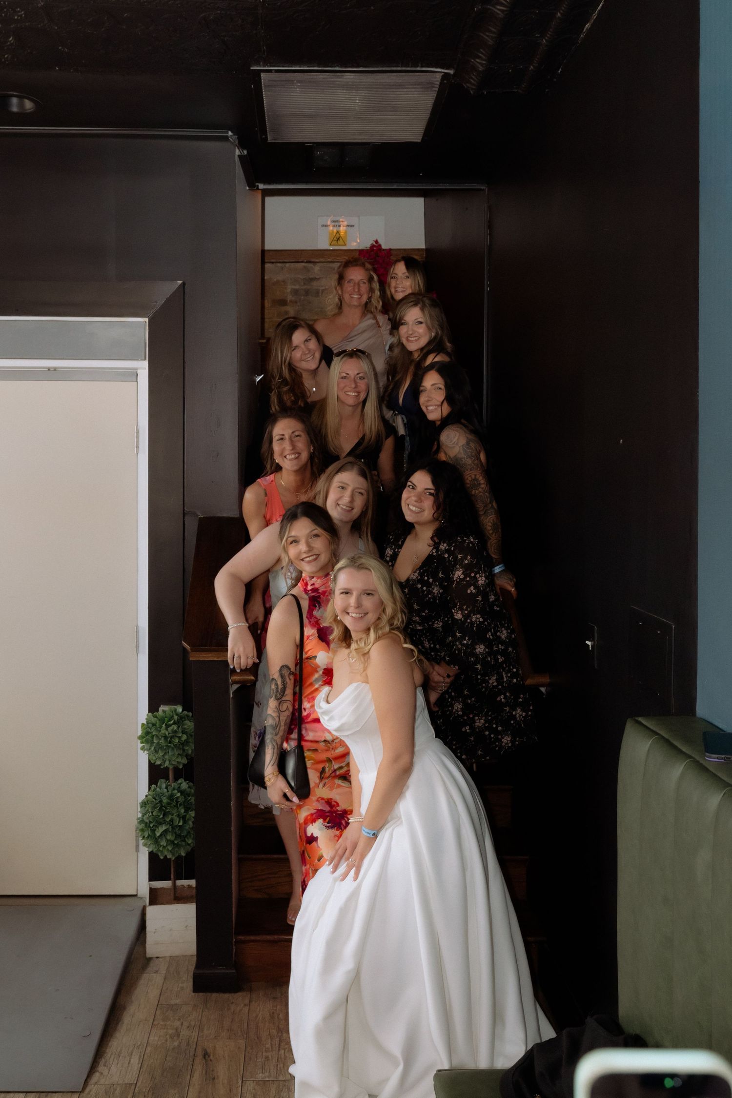 Bride in white gown poses with group of smiling women on staircase inside a dark-walled venue.