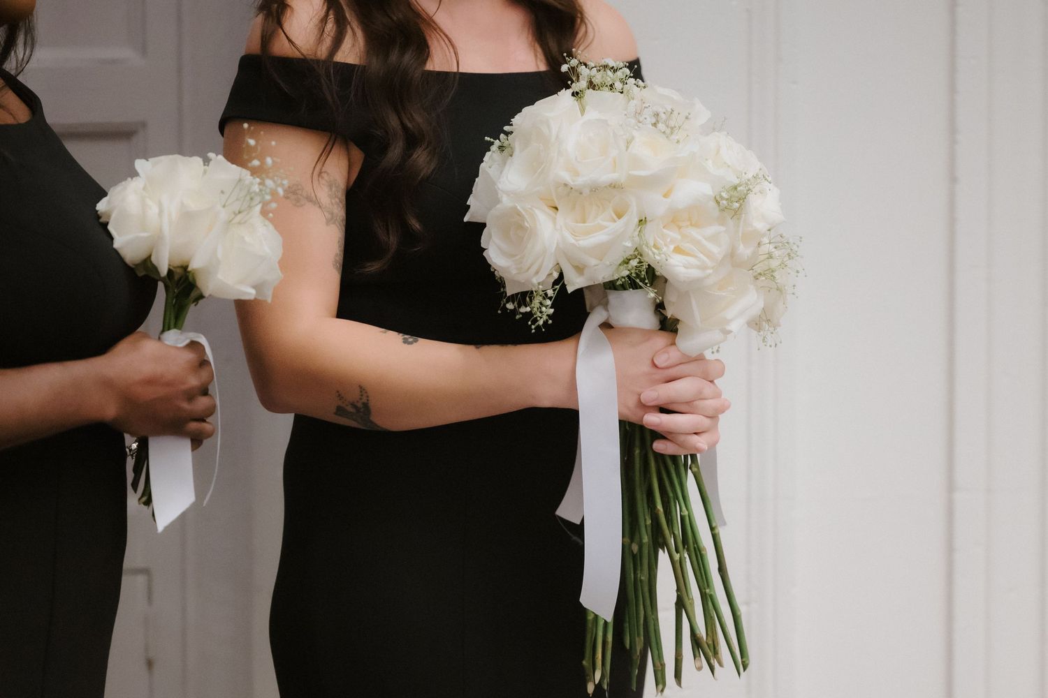 Bridesmaids in black off-shoulder dresses holding elegant white peony bouquets with white ribbon at a wedding.