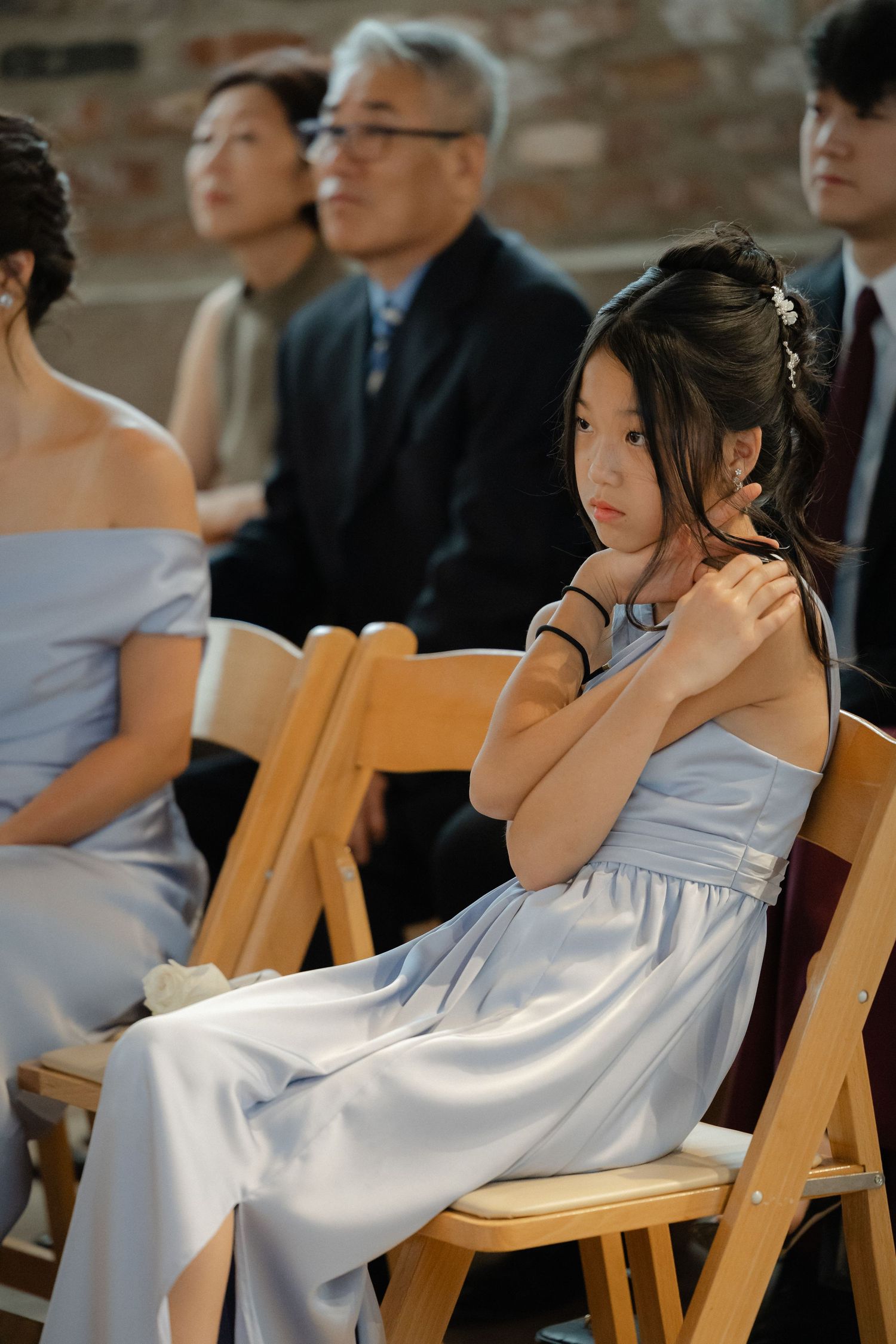 A young girl in a light blue satin dress sits attentively on a wooden folding chair during a wedding ceremony.