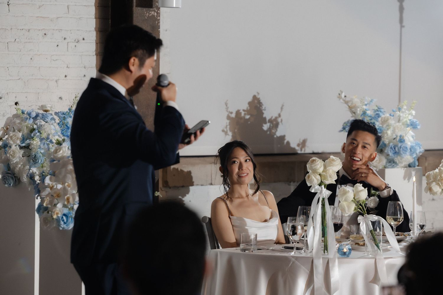 Groom giving a heartfelt wedding speech to smiling bride and groom at elegantly decorated sweetheart table.