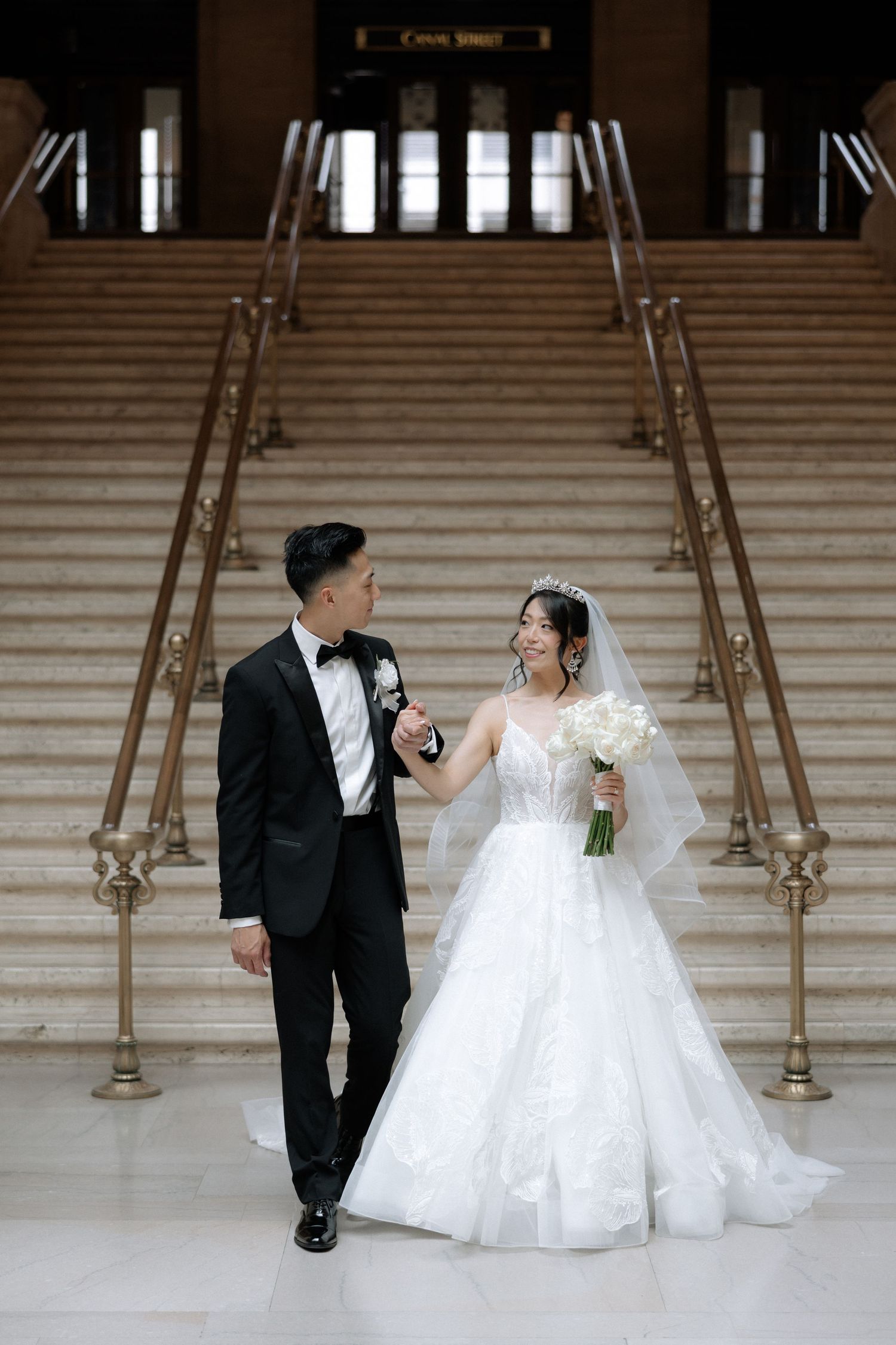 Bride and groom walk hand in hand on grand marble staircase, bride holding white bouquet in stunning wedding attire.
