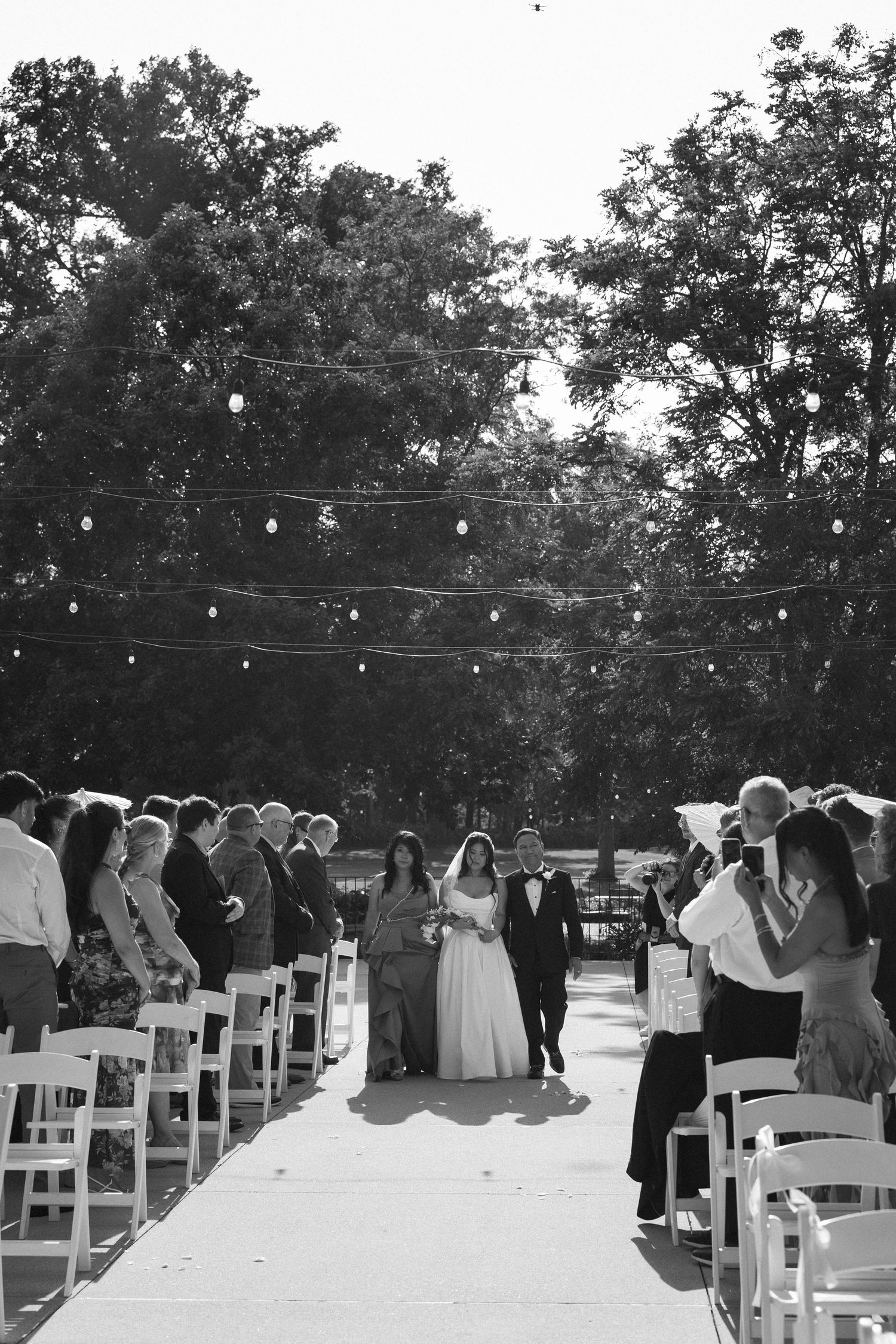 Black and white photo of bride and groom walking down outdoor wedding aisle with string lights.