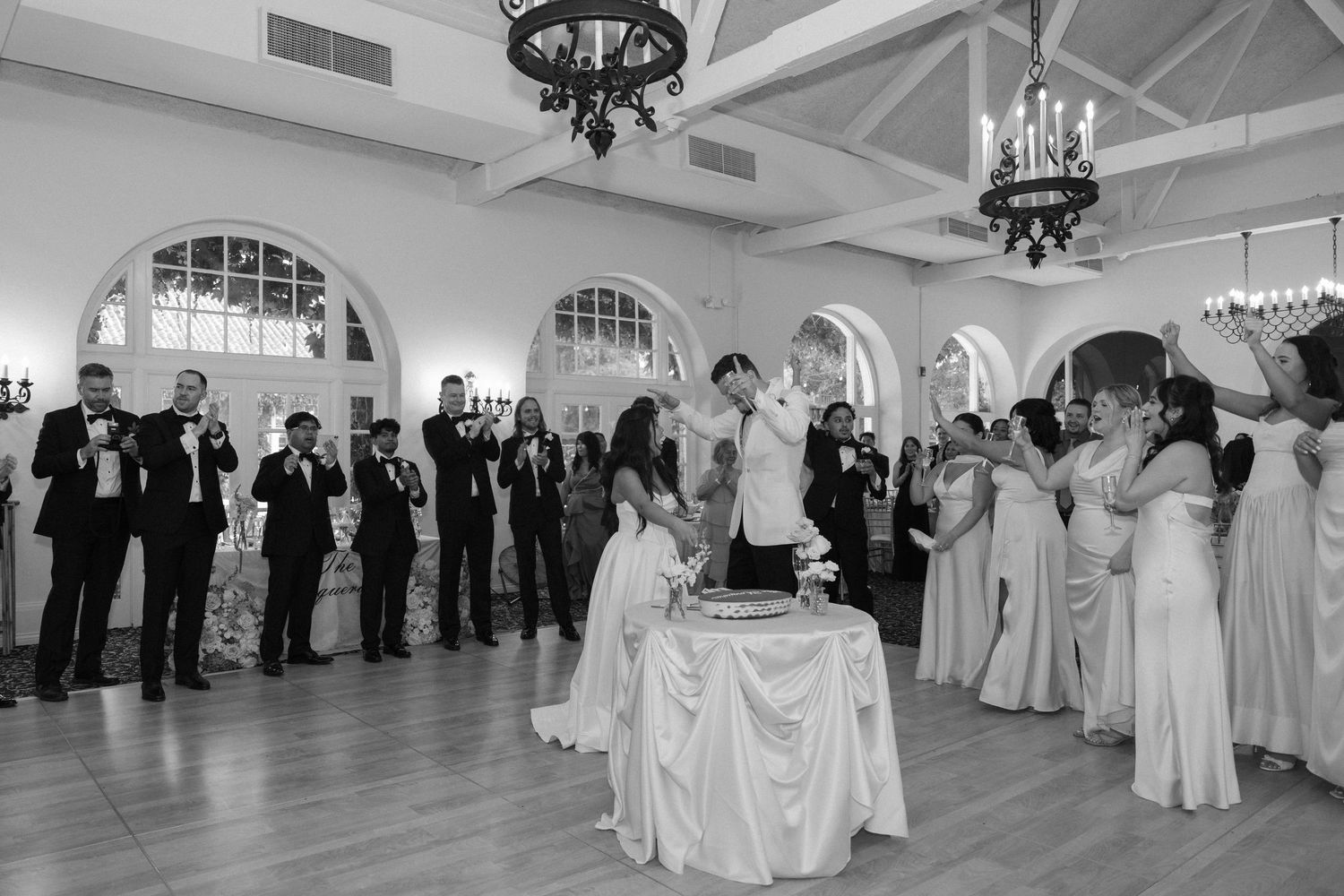 Wedding couple cuts cake while bridal party celebrates in elegant ballroom, black and white photo.