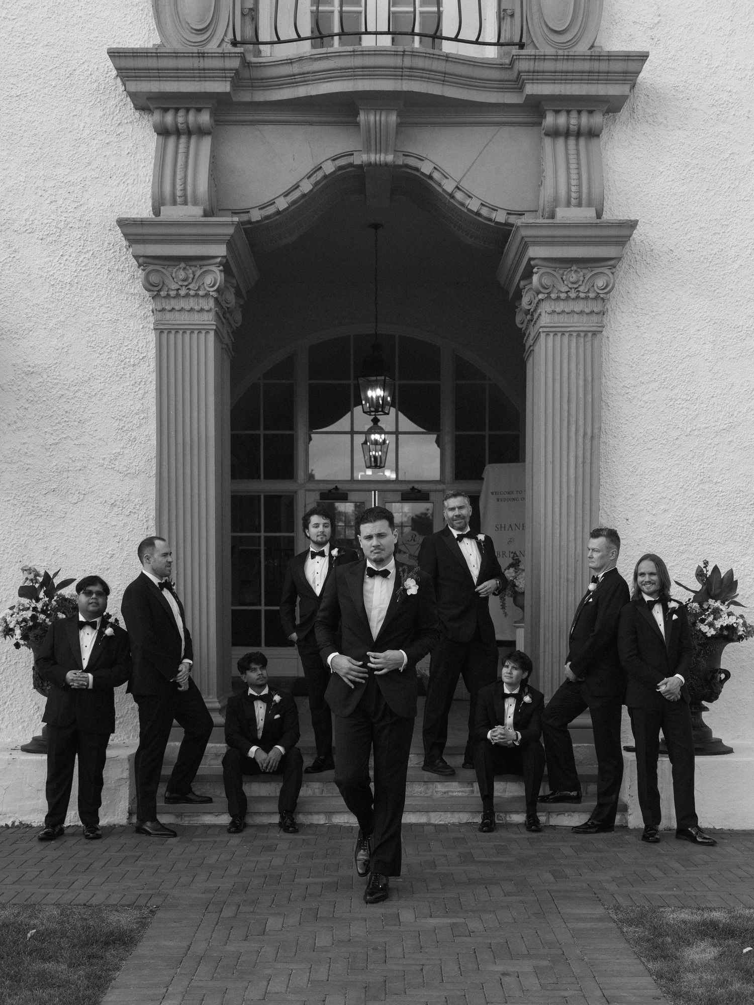 Groom leads groomsmen and ring bearers outside ornate building entrance in black and white photo.