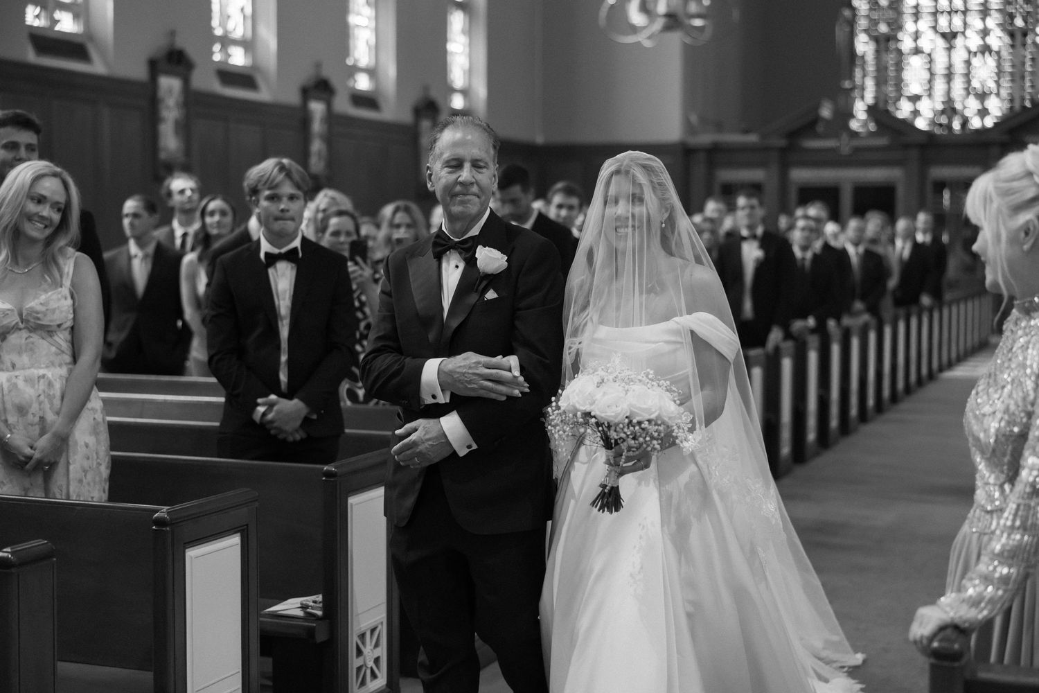 Father walks bride in white gown and veil down the church aisle in black and white wedding photo.