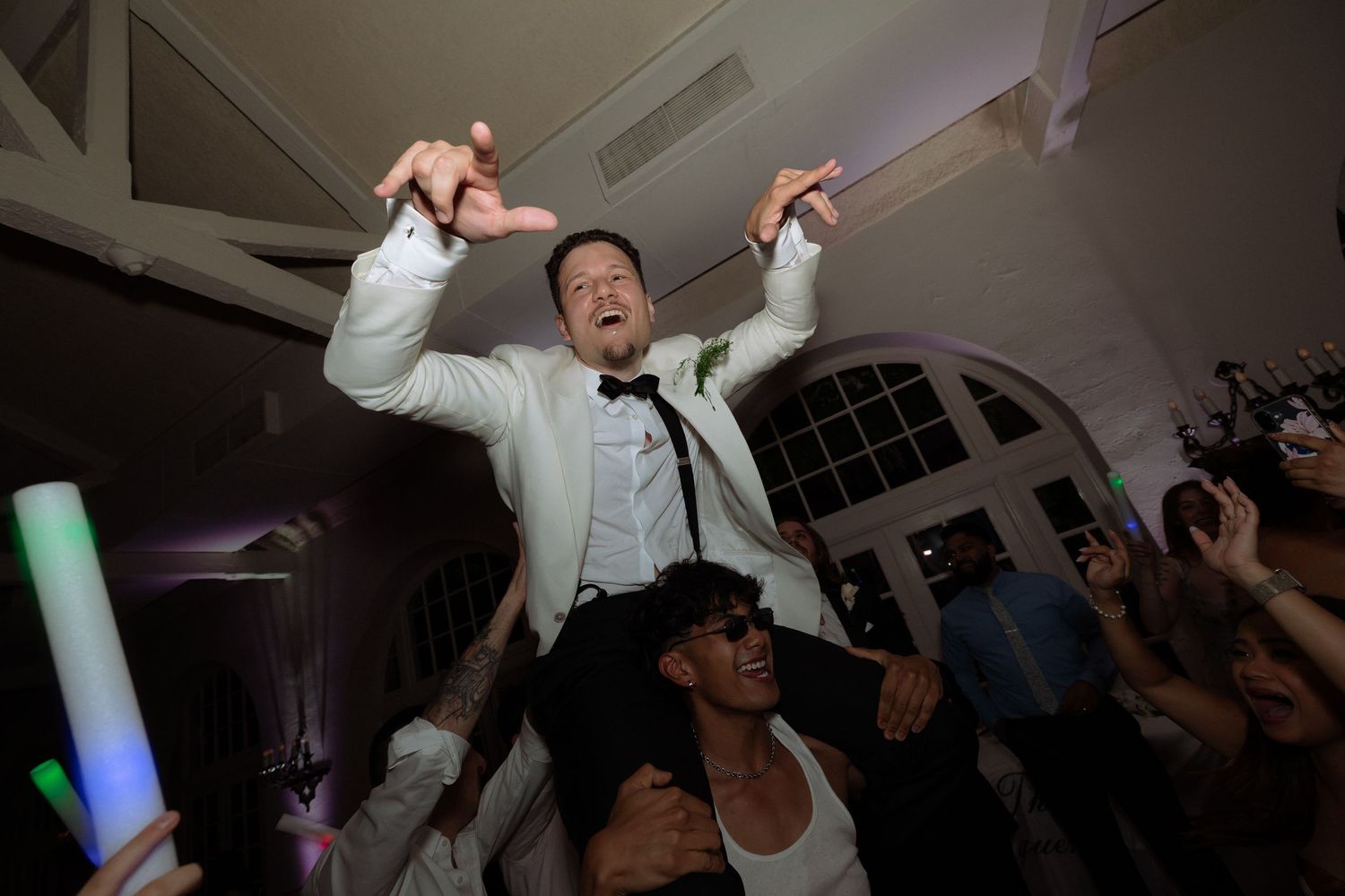 Groom being lifted on shoulders by guests during lively wedding reception dance floor celebration.