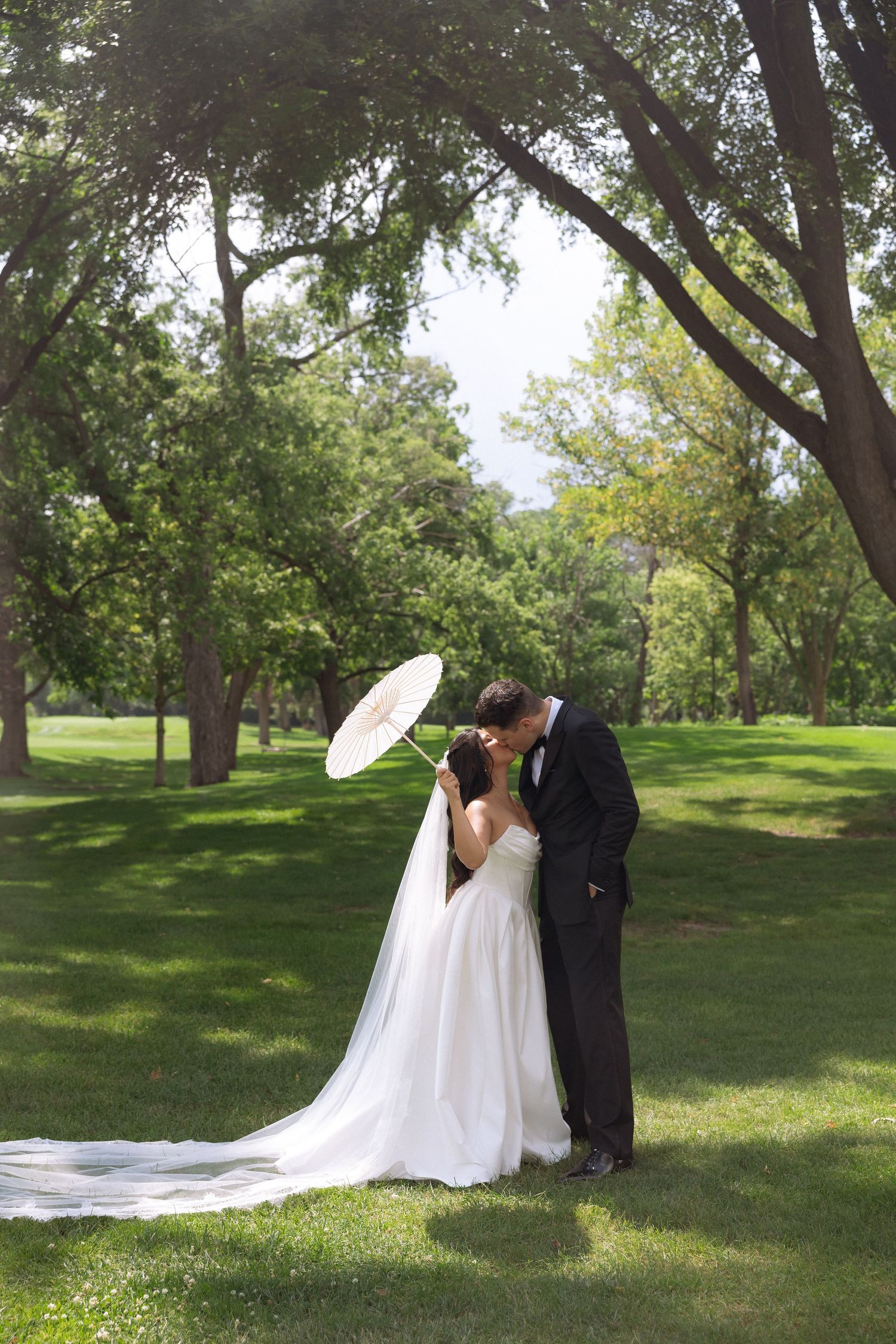 Bride and groom kiss under white parasol umbrella surrounded by lush green trees in sunny park.