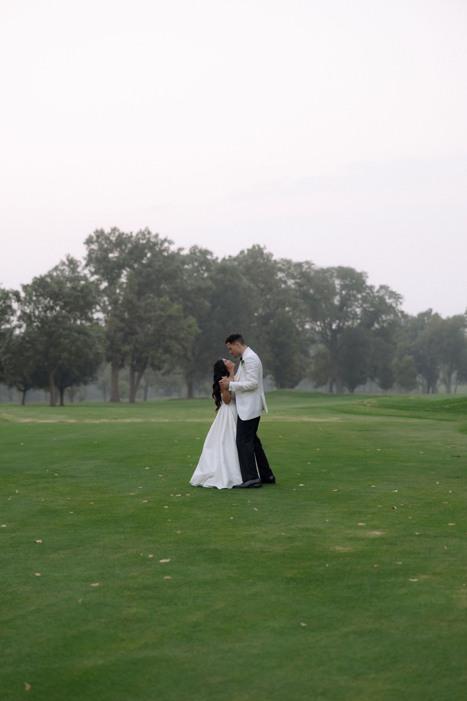 Bride and groom embrace romantically on a lush green golf course surrounded by misty trees.