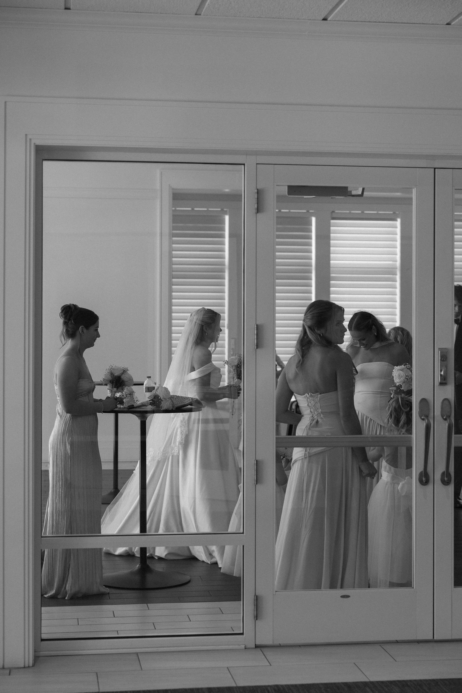 Black and white photo of bride with bridesmaids preparing inside venue viewed through glass French doors.