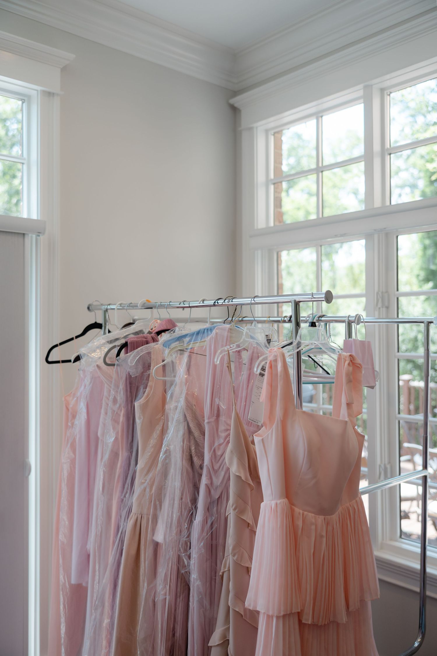 Pink bridesmaid dresses hanging on a silver garment rack near bright white windows in an elegant bridal suite.