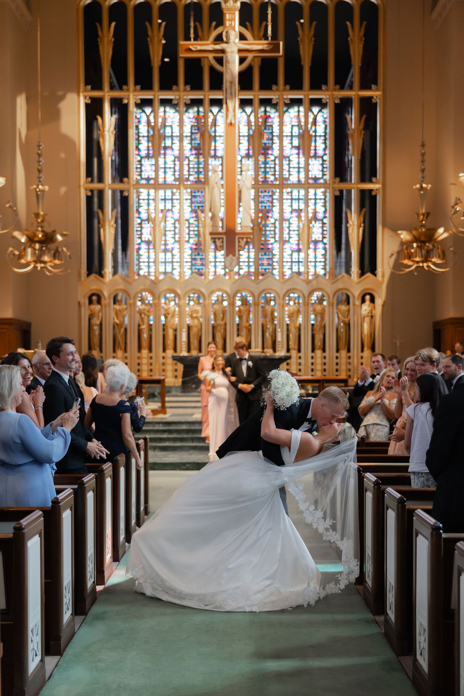 Groom dips bride in white gown for a kiss in church aisle as guests applaud their union.