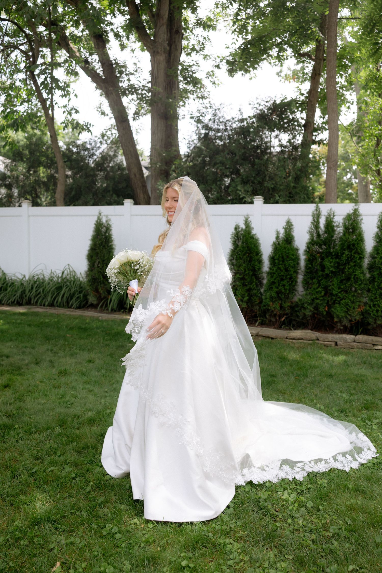 Elegant bride in white ball gown with lace veil holding bouquet standing on green lawn outdoors.