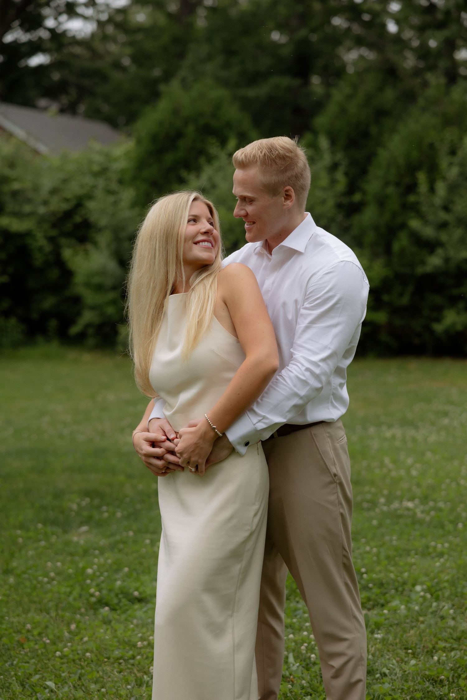 A couple embraces in a lush green garden, the woman in a white dress and man in a white shirt and khaki pants.