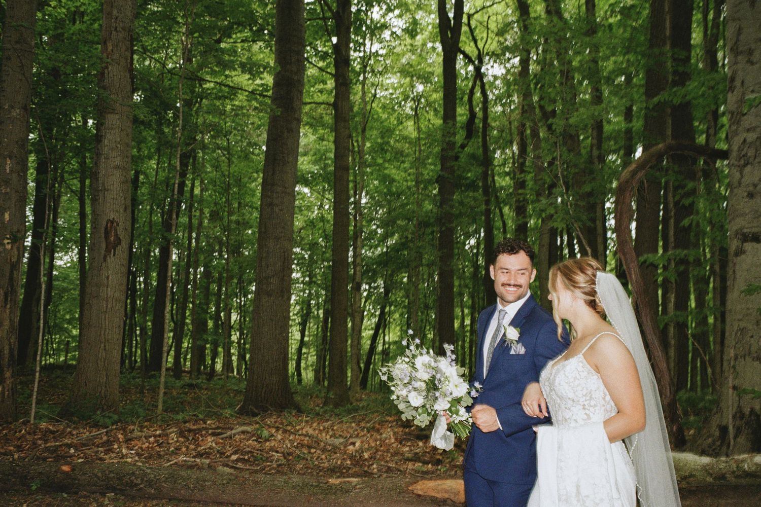 Bride and groom pose together in a lush green forest, bride holding white bouquet, groom in blue suit.