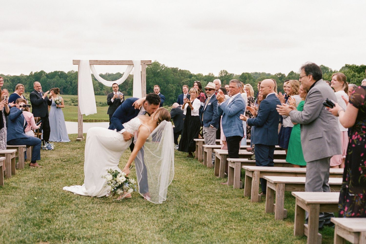 Bride and groom share a romantic dip kiss at outdoor wedding ceremony with wooden arch and seated guests.