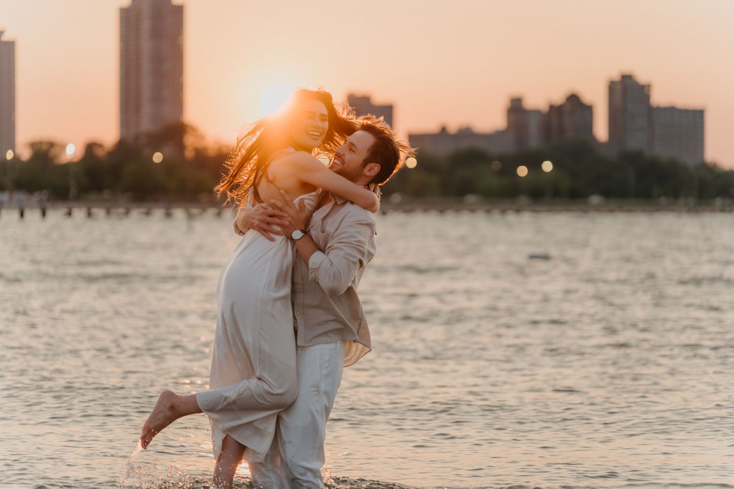 Couple embraces on beach at golden sunset with city skyline reflecting on calm water behind them.