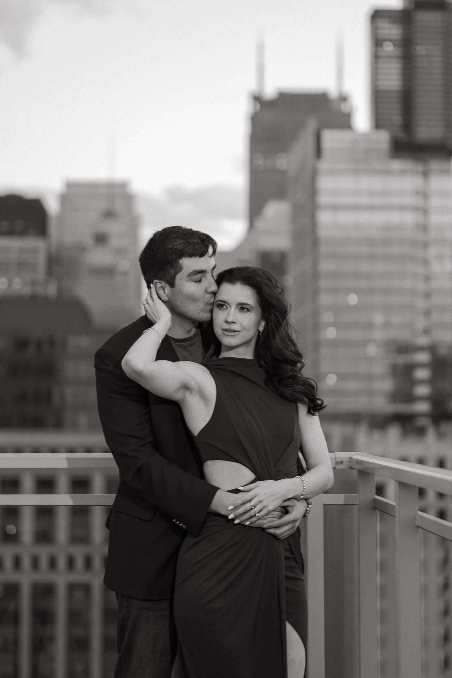 Black and white romantic photo of man kissing woman on cheek on urban rooftop.