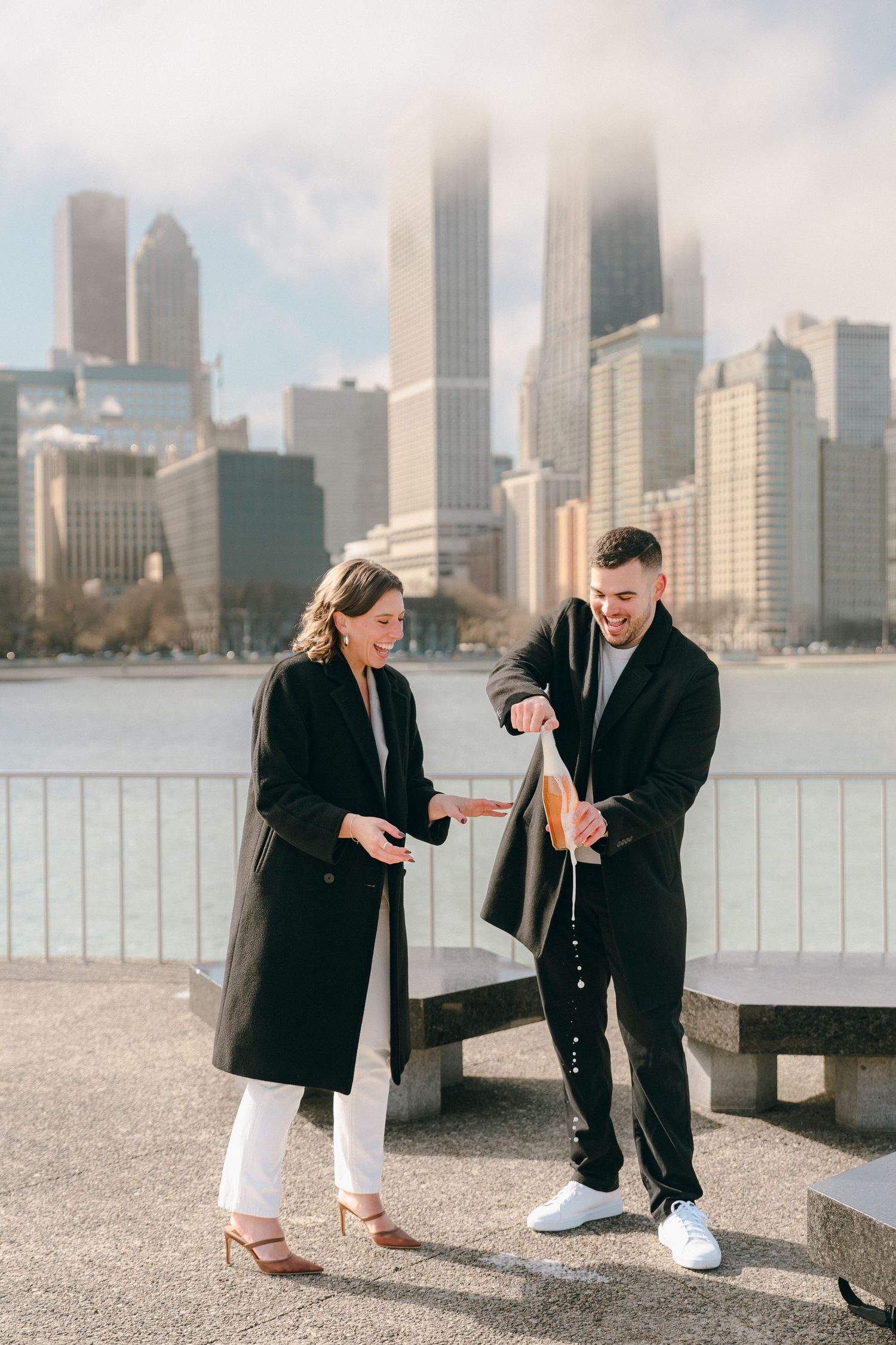 Couple in black coats opening champagne bottle by Chicago waterfront skyline on sunny day.