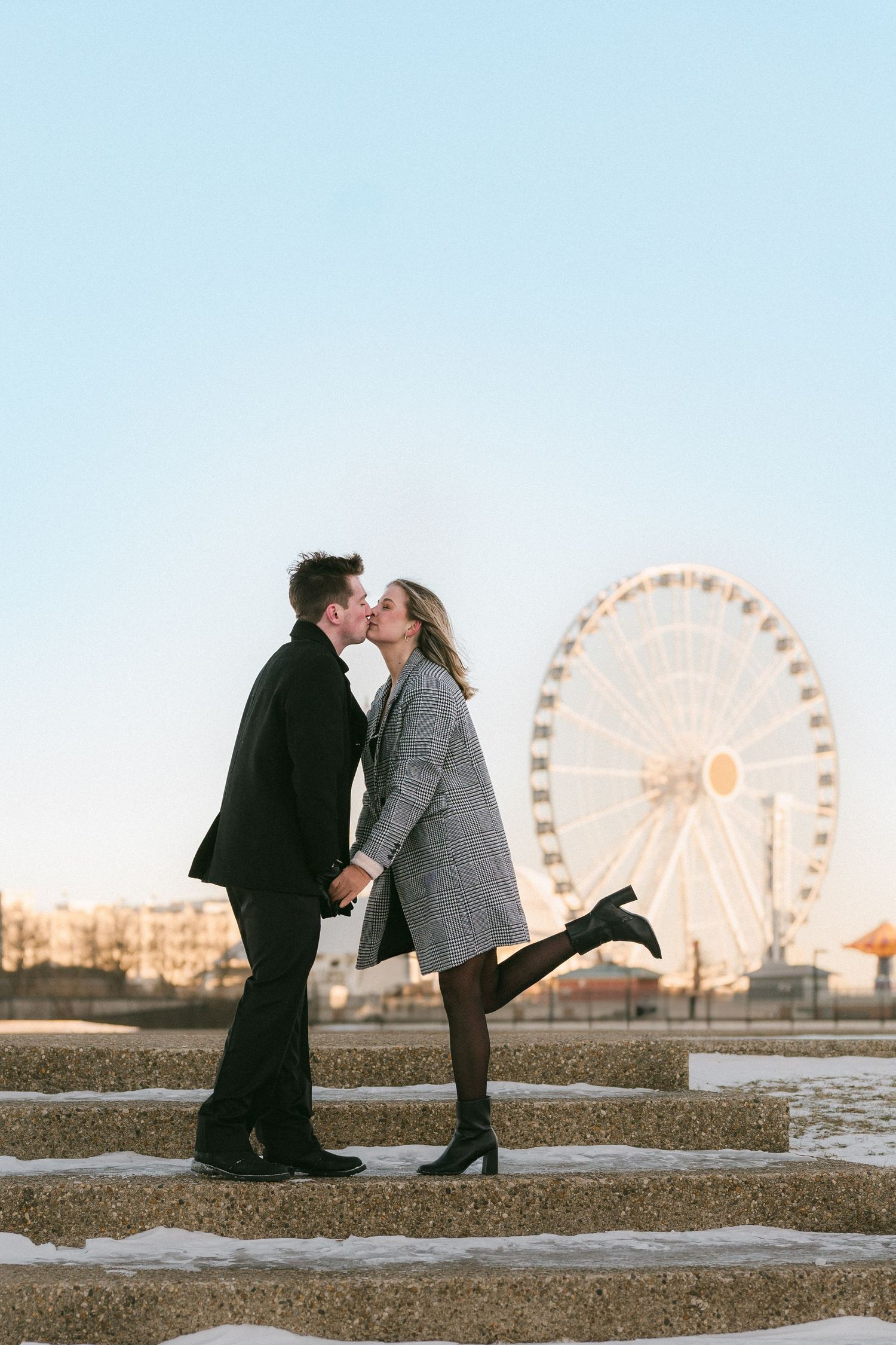 A couple shares a romantic kiss on stone steps with a Ferris wheel glowing in the background.