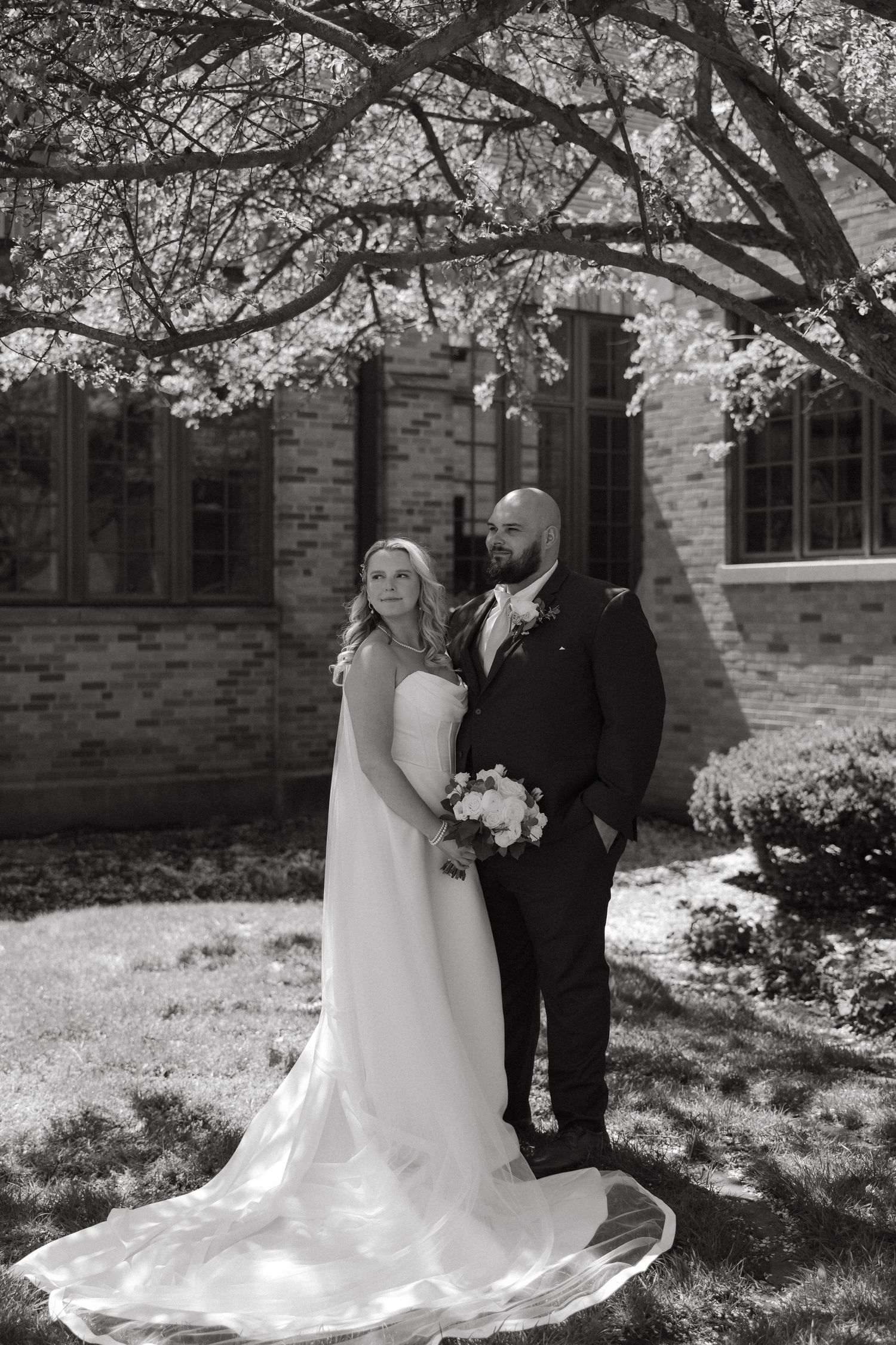 Black and white wedding photo of bride in flowing gown and groom in suit under blooming tree by brick church.