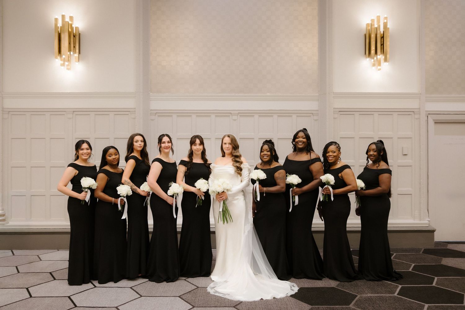 Bride in white gown poses with ten bridesmaids in black dresses holding white bouquets in elegant ballroom.