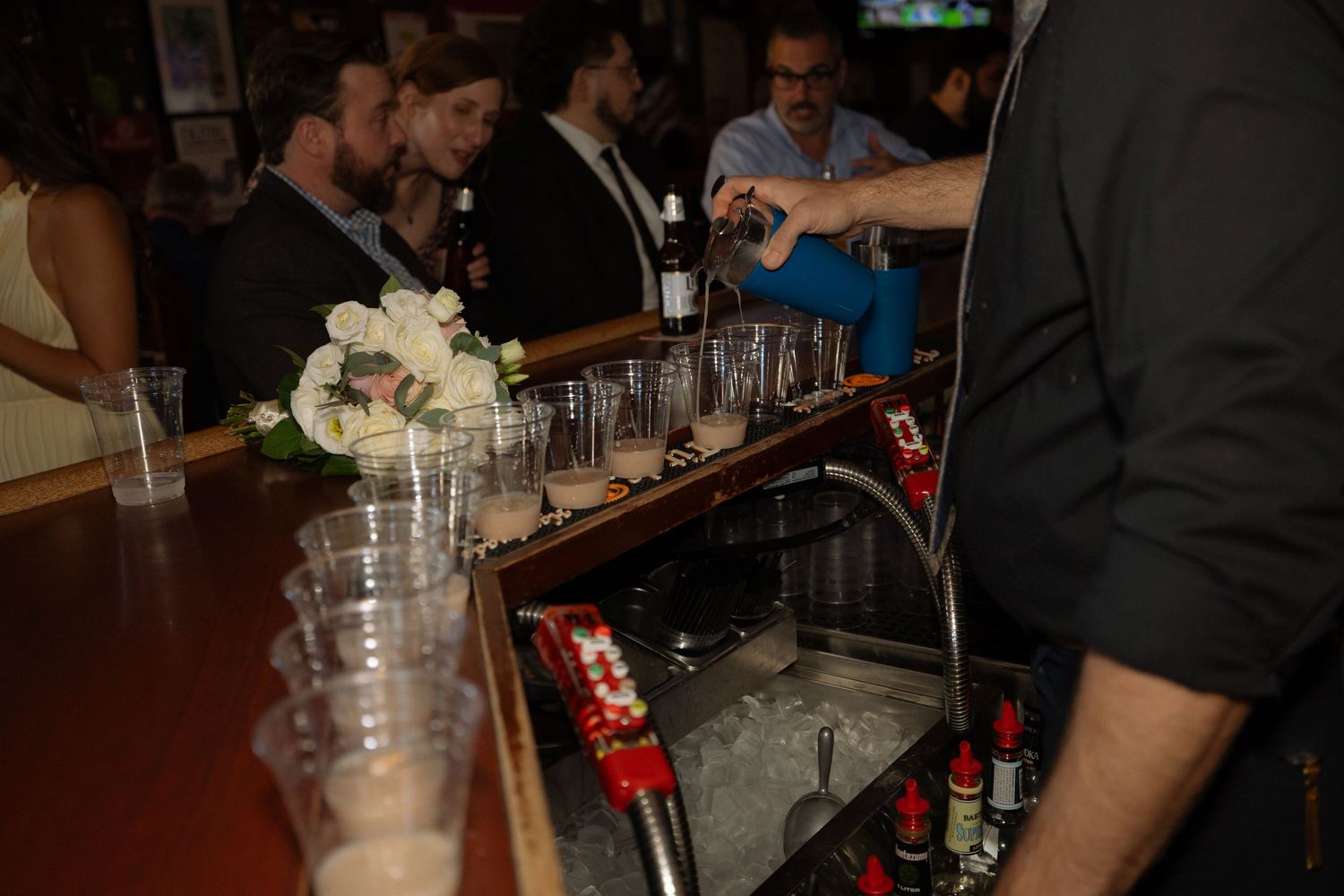 Bartender pouring drinks at a busy bar with white floral bouquet and guests in background.