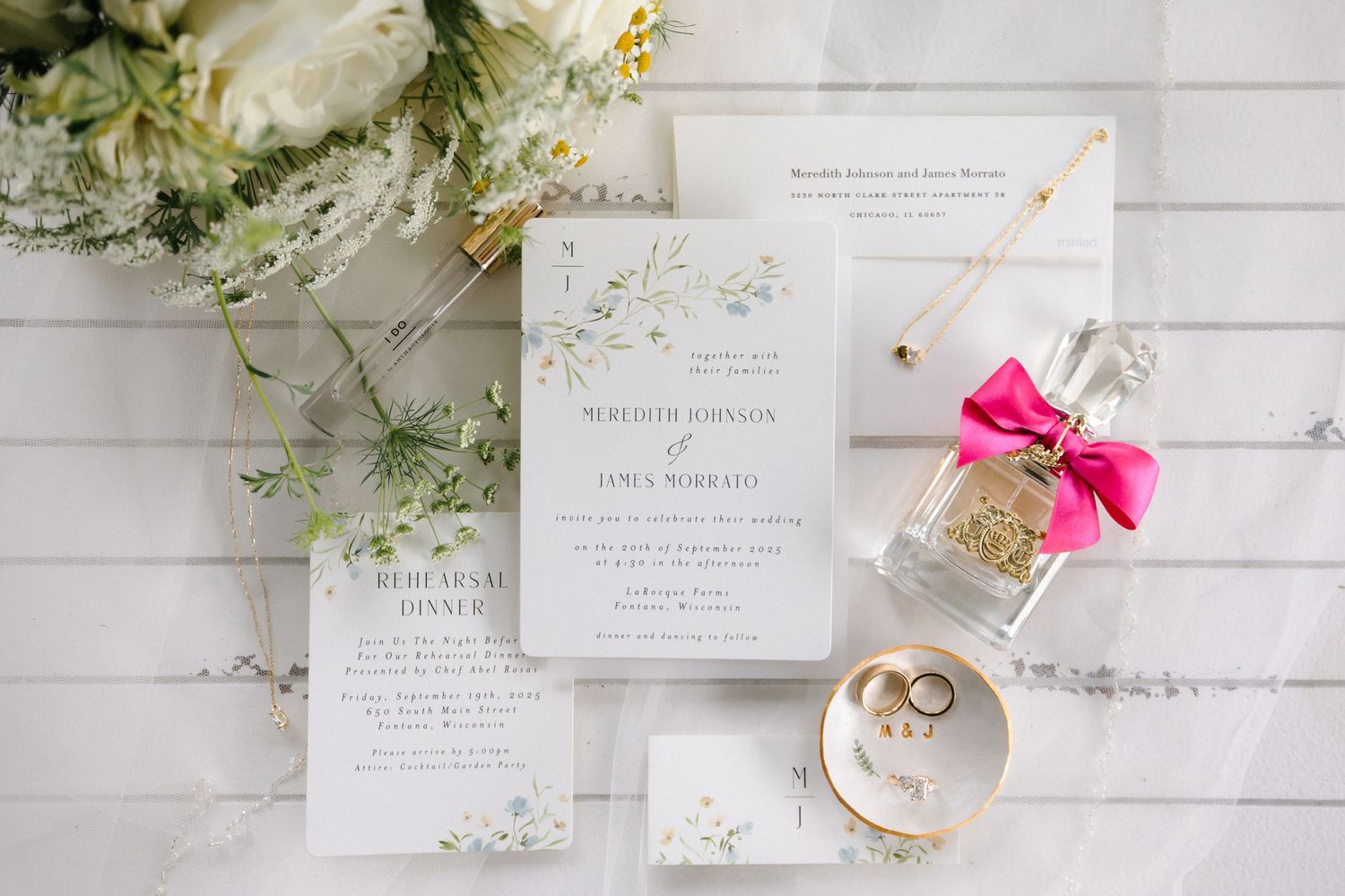 Elegant wedding invitation suite with greenery details displayed with rings, perfume bottle, and white floral bouquet.