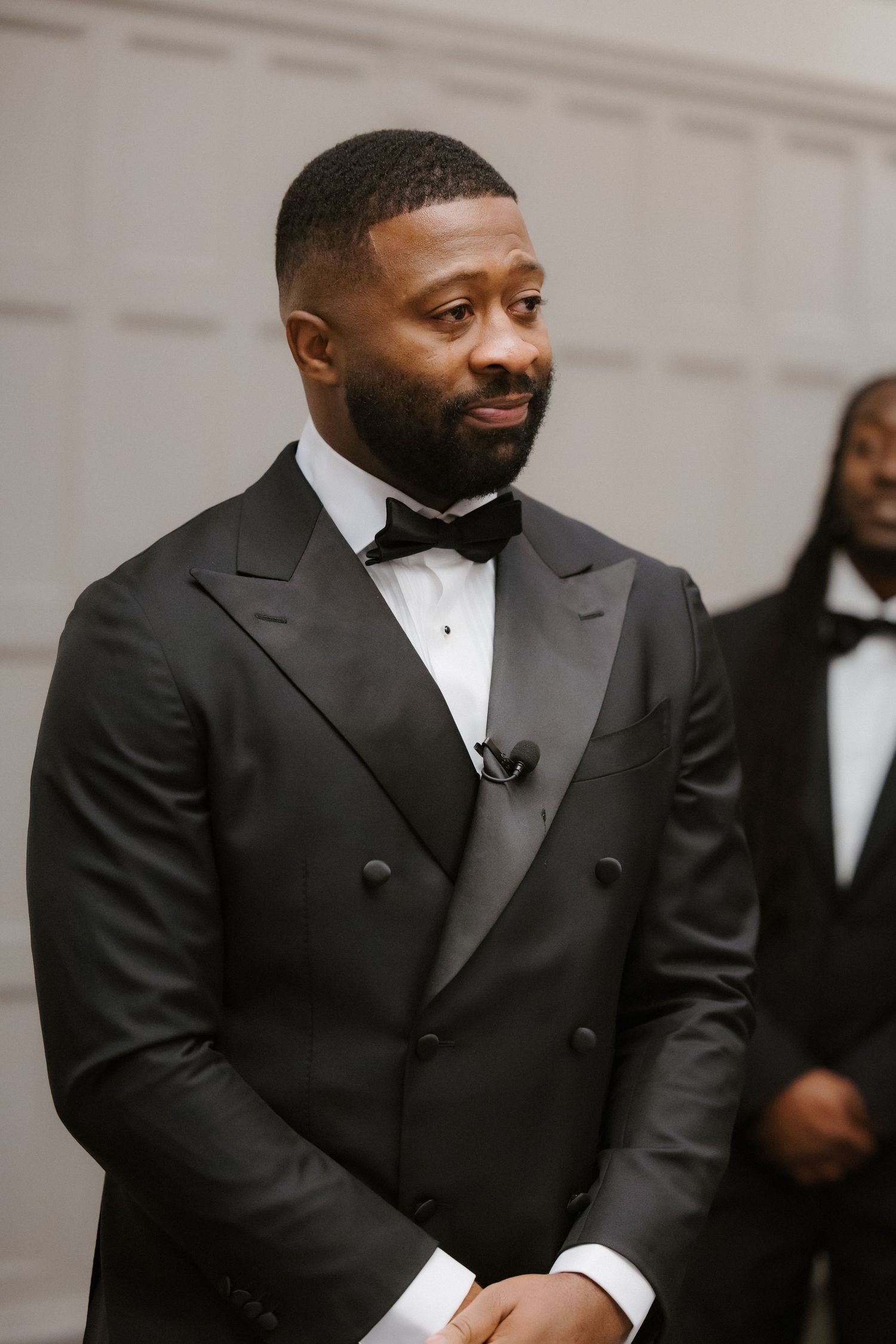 A man in a black double-breasted tuxedo with bow tie stands elegantly at a formal event.