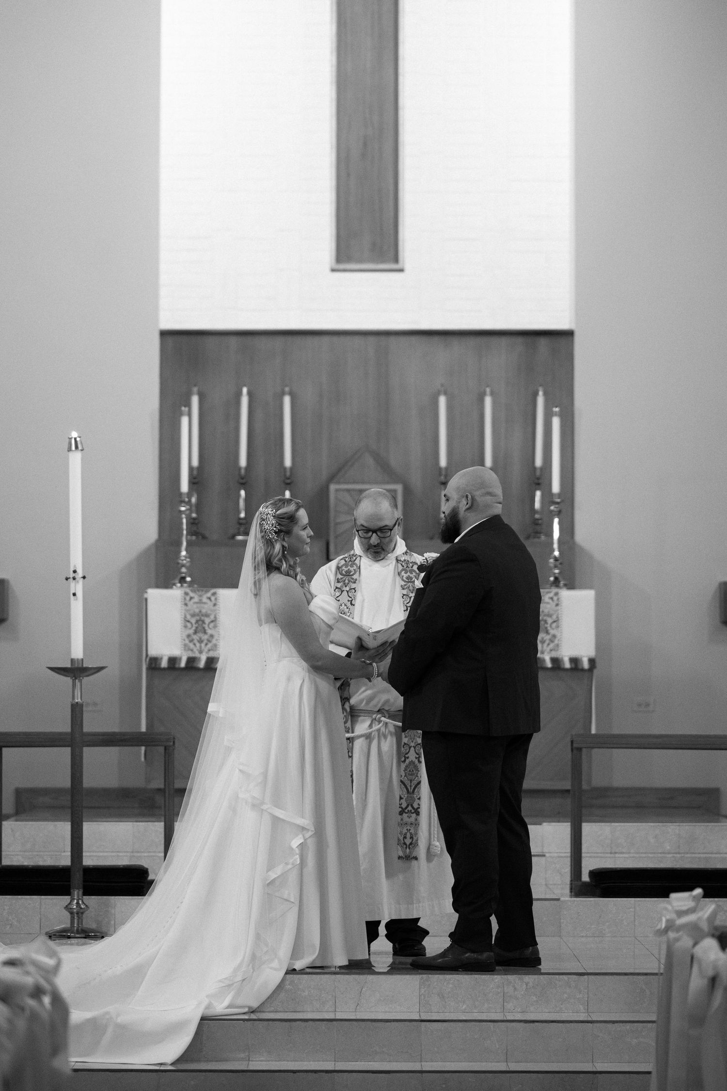 Black and white photo of a bride and groom exchanging vows at a church altar with a officiant.