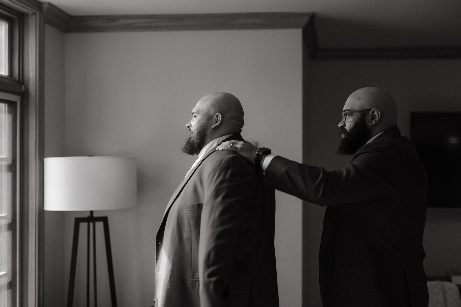 Black and white photo of a man helping another man adjust his suit jacket before a wedding ceremony.