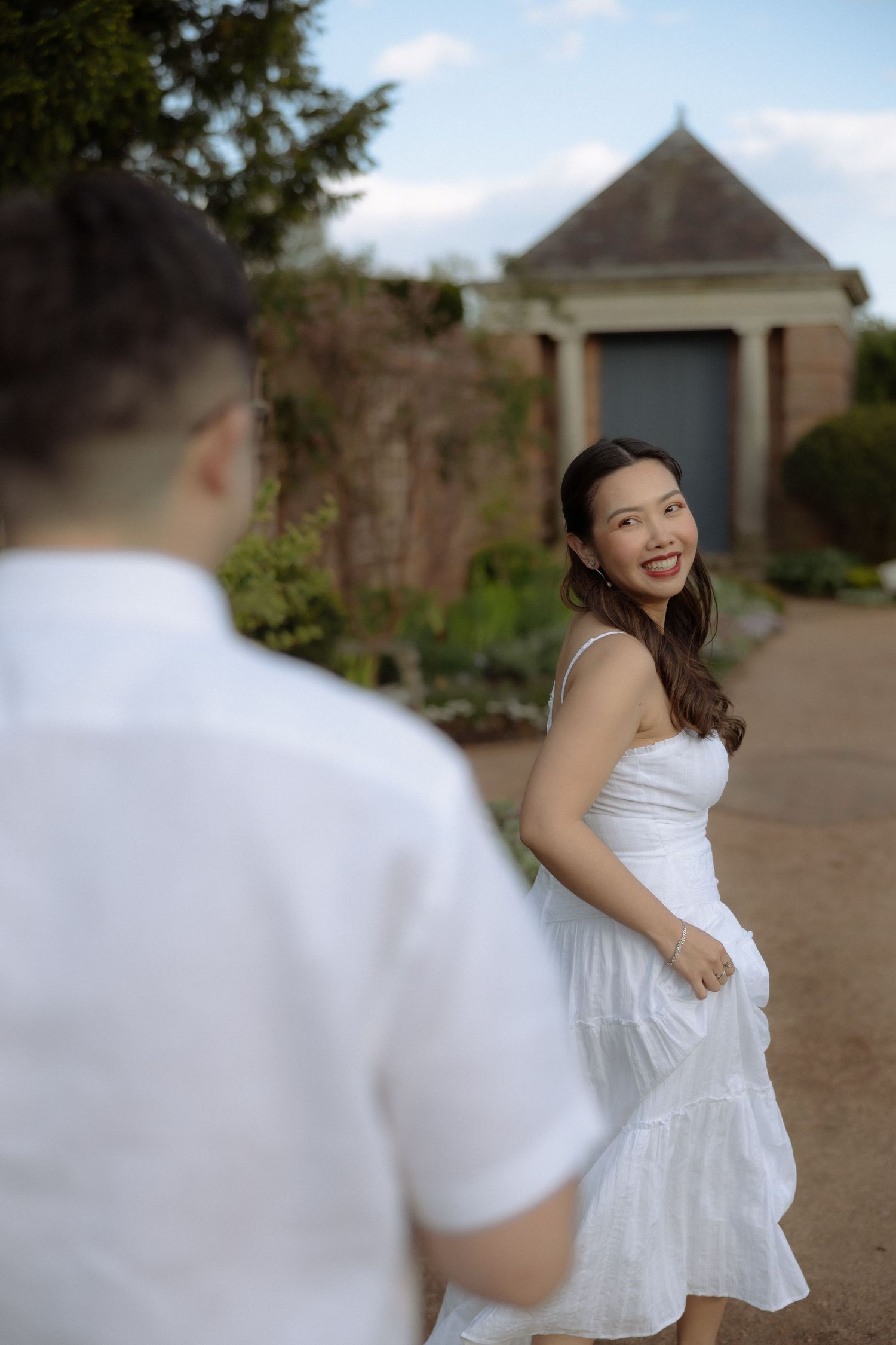 Smiling Asian woman in white dress looks back at man in garden setting near brick building.