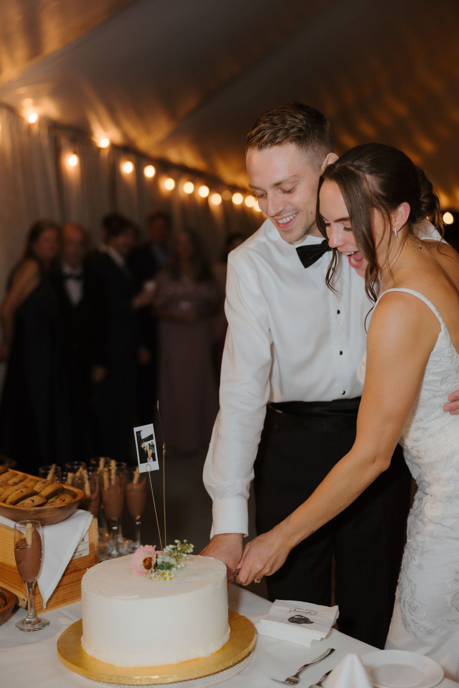 Smiling newlyweds cut their white wedding cake together under warm tent lighting at their reception.