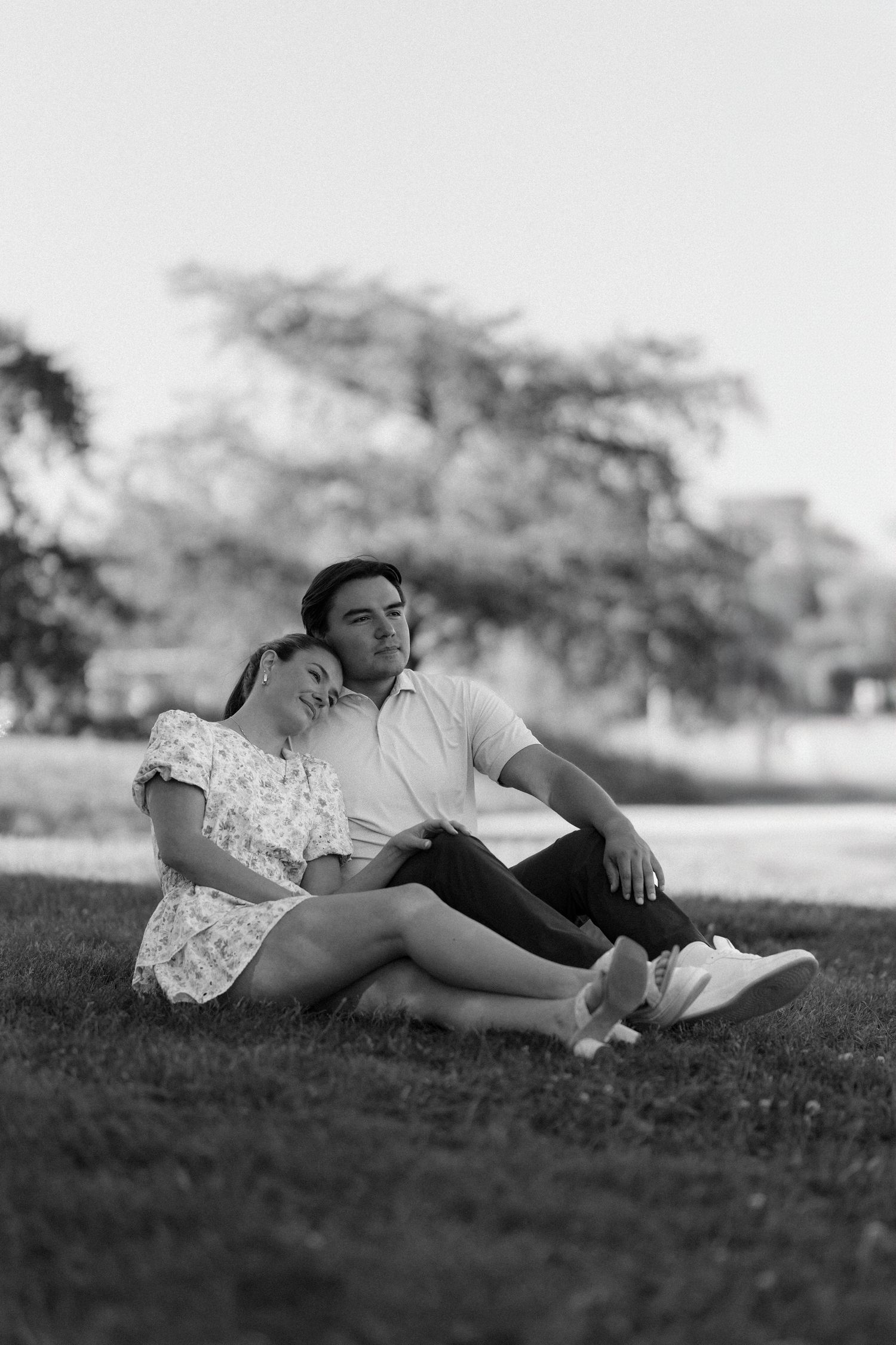 Black and white photo of couple sitting together on grass leaning on each other in peaceful park setting.