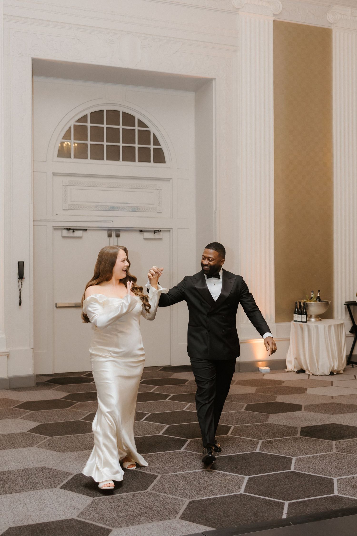 A bride and groom share their first dance in an elegant ballroom, she in a white satin gown, he in a black tuxedo.