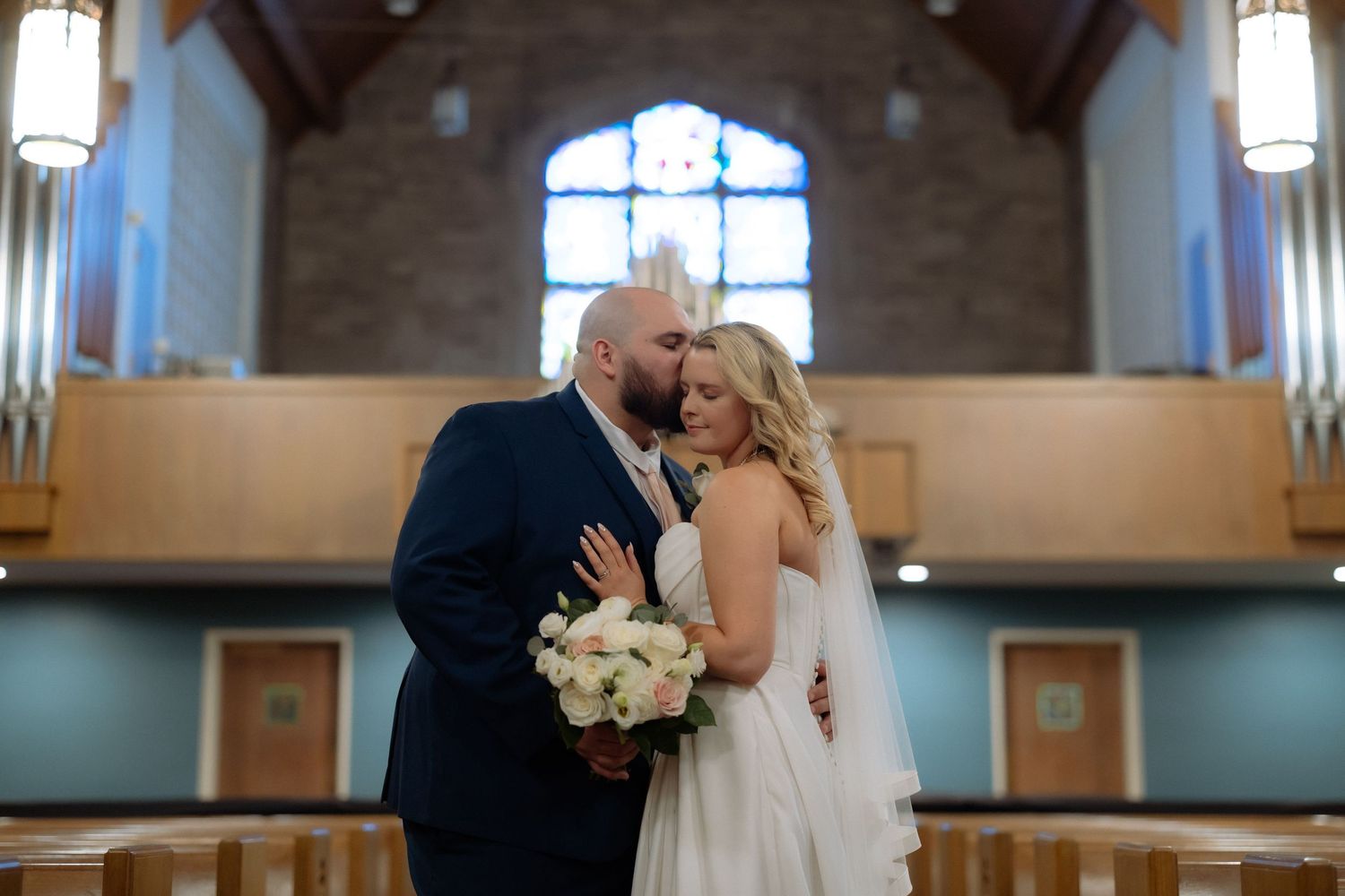 Bride and groom share a romantic kiss inside a church, bride holding white rose bouquet, stained glass window behind them.