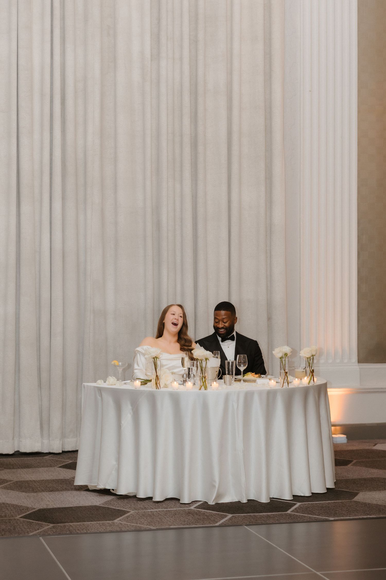 Bride and groom laughing at sweetheart table with white linens and gold floral accents at elegant wedding reception.