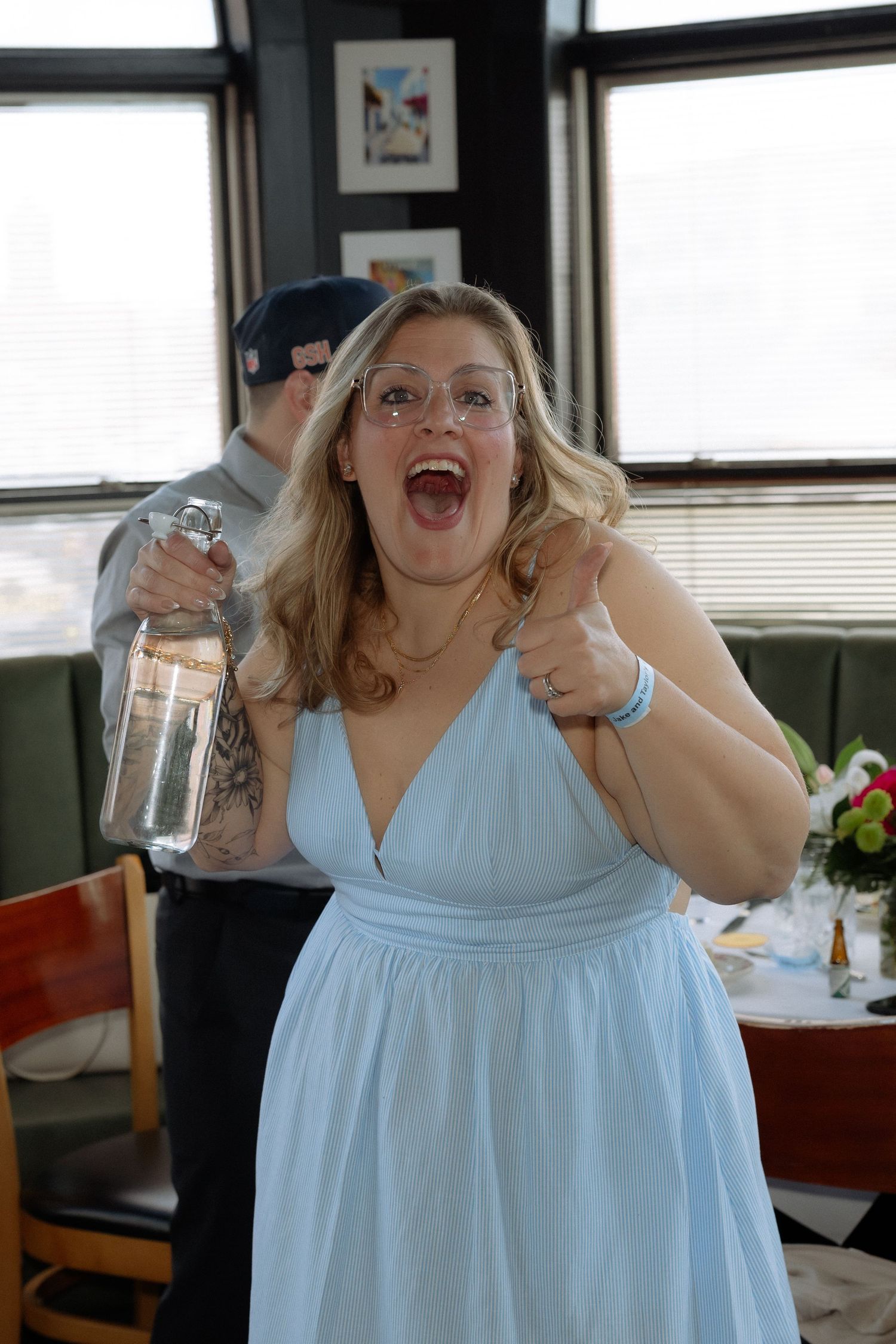Excited blonde woman in light blue dress holds bottle and gives thumbs up at indoor wedding reception.