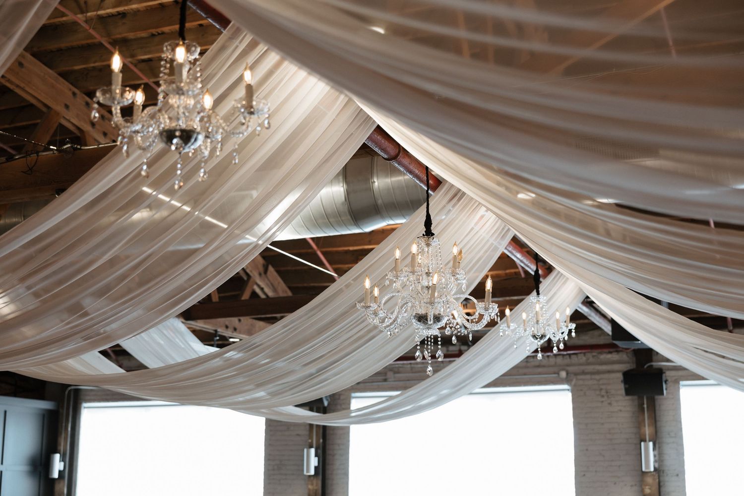 Elegant event venue ceiling draped with sheer white fabric and crystal chandeliers for a wedding reception.