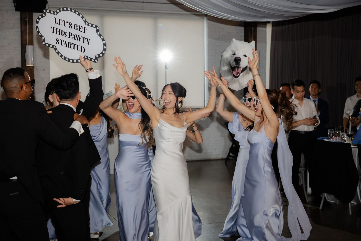 Bridesmaids in blue dresses cheer with bride during wedding reception entrance with polar bear mascot.